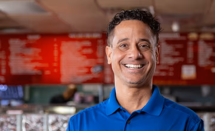 Smiling man in a blue polo shirt standing in front of a blurred red menu board inside a restaurant.
