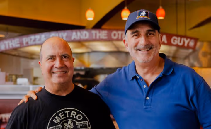 Two smiling men standing closely together inside a pizzeria, one wearing a black Metro t-shirt and the other a blue polo shirt and a cap.