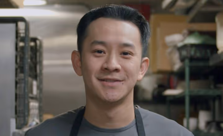 Smiling man with short black hair wearing a gray shirt and black apron in a kitchen setting.