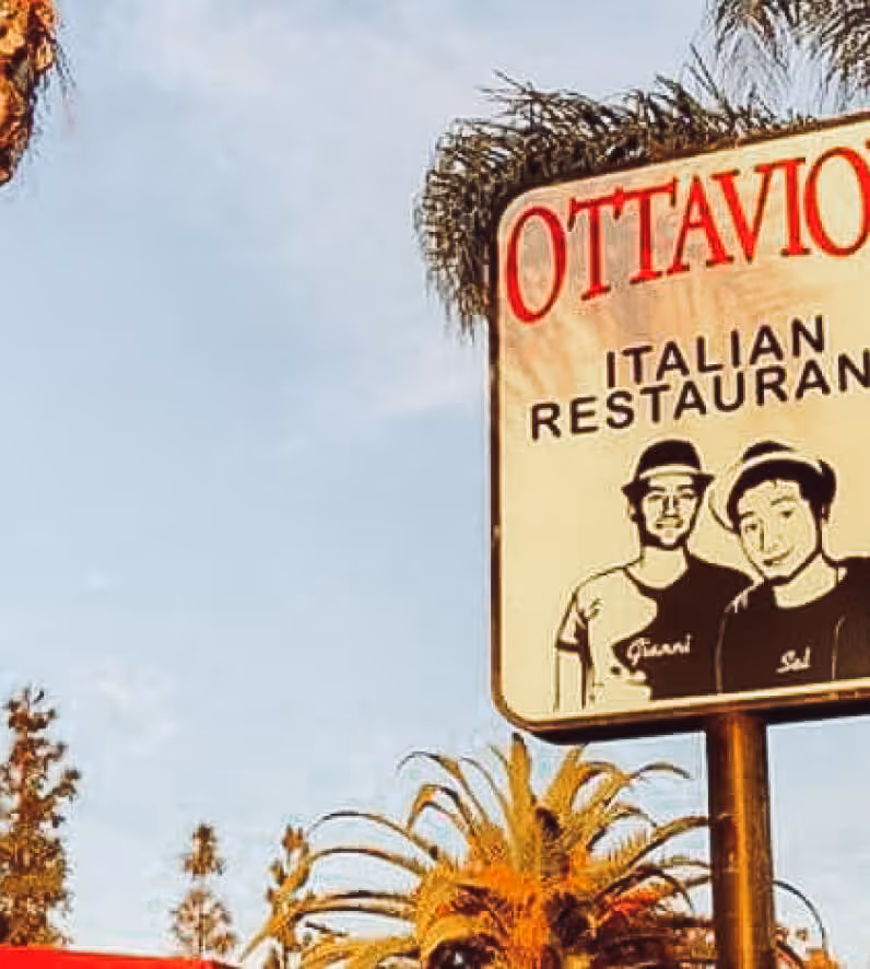 Sign for Ottavio Italian Restaurant with illustrations of two men wearing hats, surrounded by palm trees and blue sky.