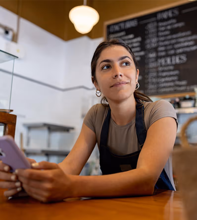 Young woman in a cafe apron sitting at a wooden table, holding a smartphone and looking thoughtfully to the side.