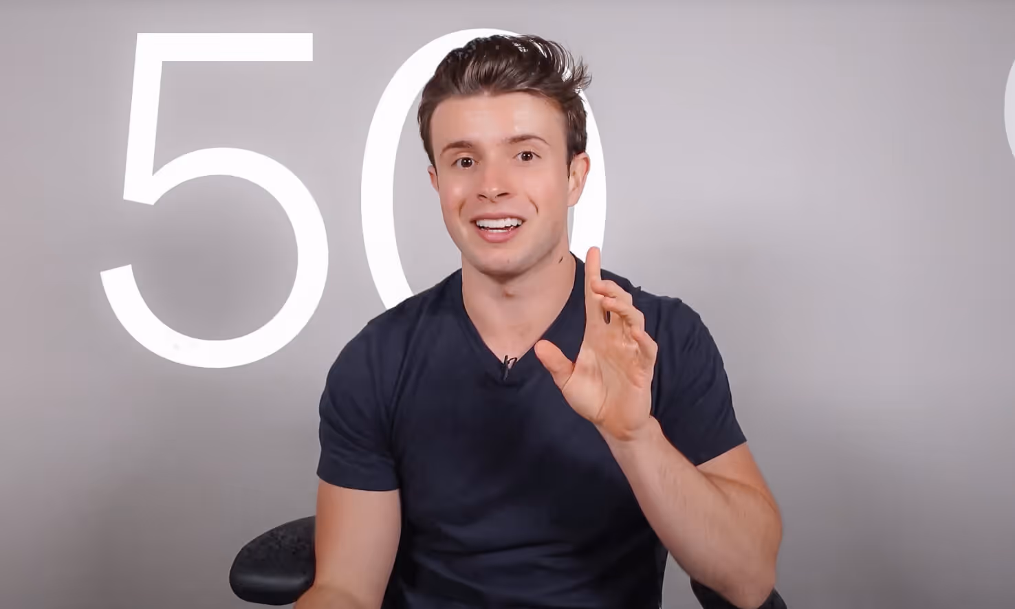 Smiling young man in a black t-shirt speaking with hand raised against a gray background with large white number 50.