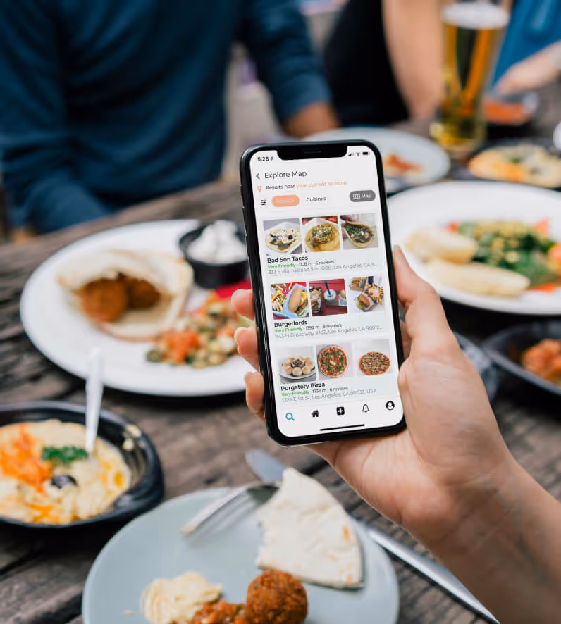 Hand holding a smartphone showing a food restaurant app with listings for tacos, burgers, and pizza, with plates of food and drinks on a wooden table in the background.