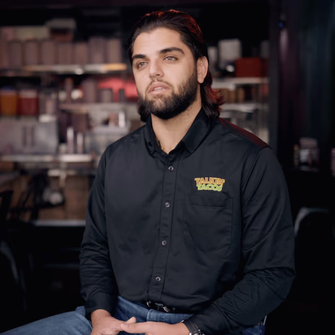 Man with a beard wearing a black Talkin' Tacos shirt sitting indoors in a casual setting.