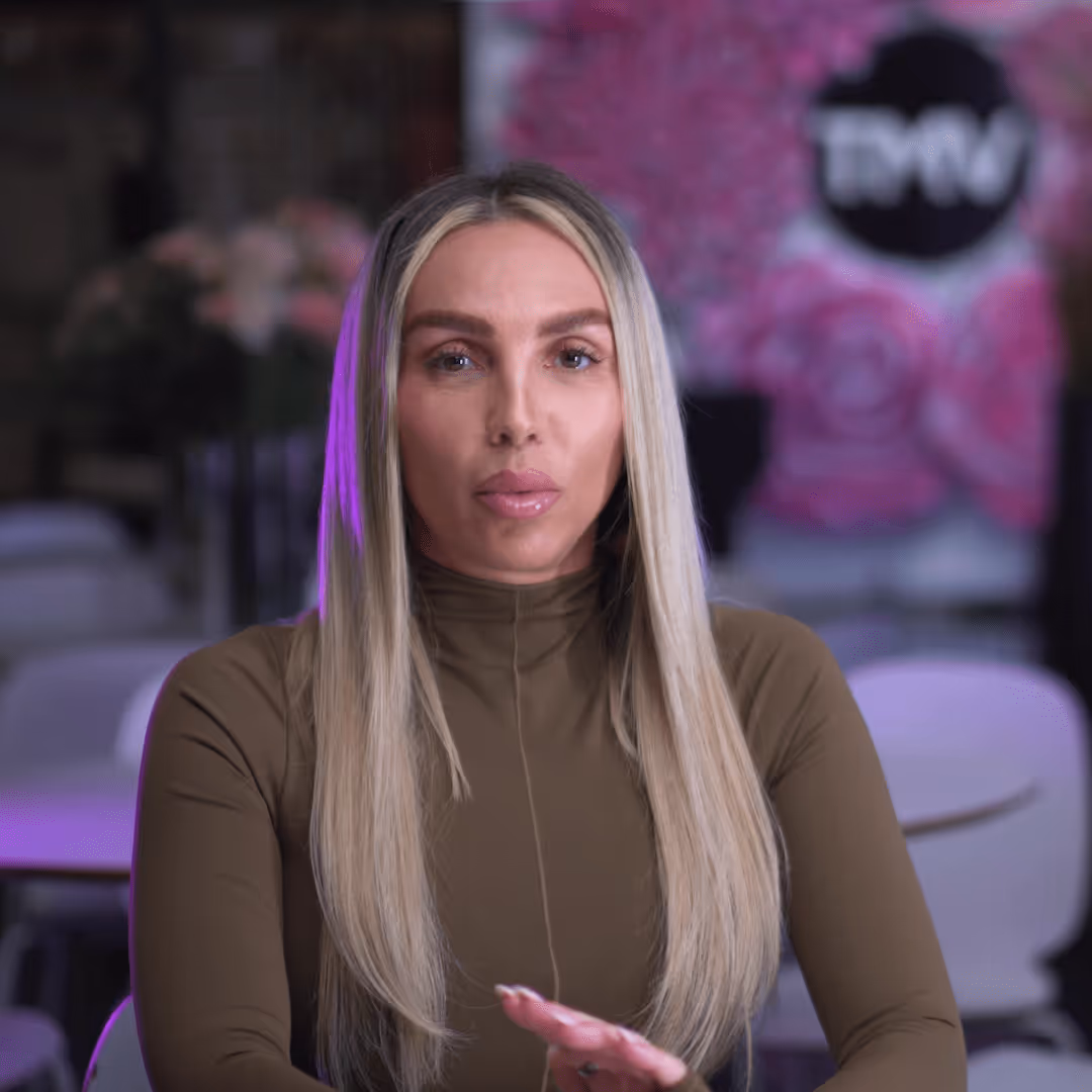 Woman with long blonde hair wearing a brown turtleneck, sitting indoors with a blurred background of pink flowers and a TVN logo.
