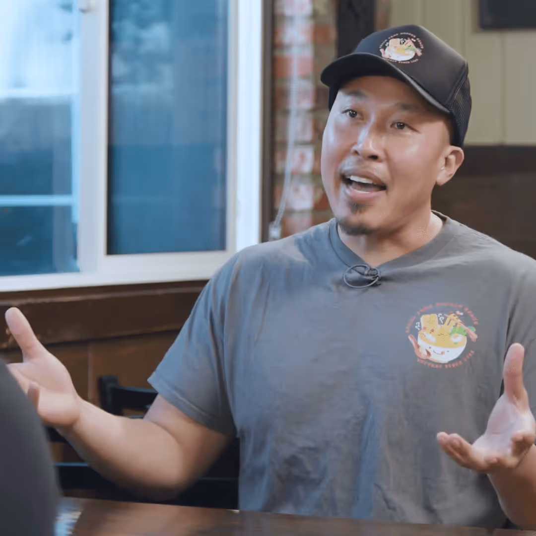 Man wearing a black cap and gray t-shirt with a noodle bowl logo, speaking and gesturing with hands at a wooden table in a casual dining setting.