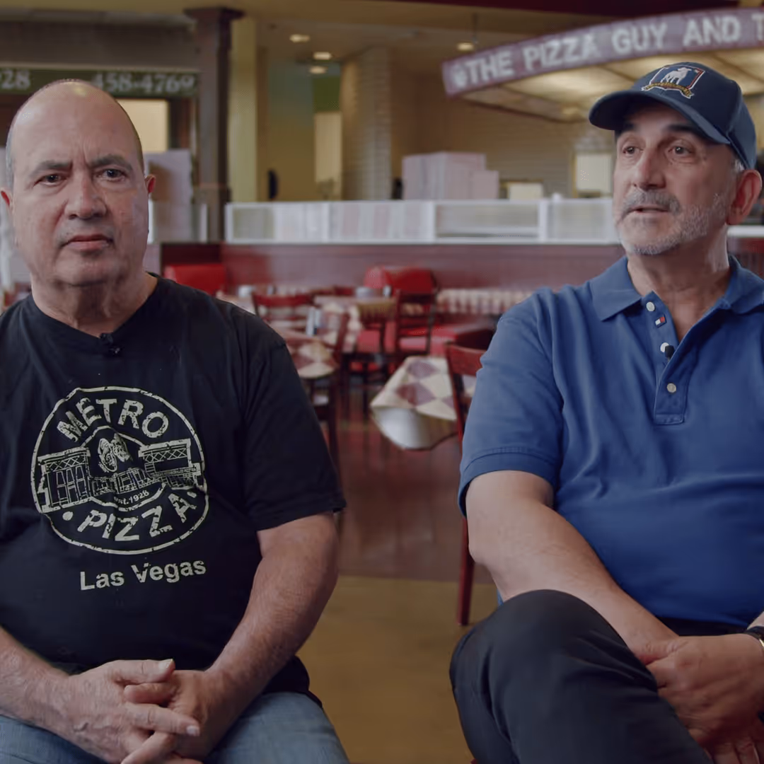 Two men sitting in a restaurant interior with empty tables and chairs, one wearing a Metro Pizza Las Vegas t-shirt and the other a blue polo and cap.