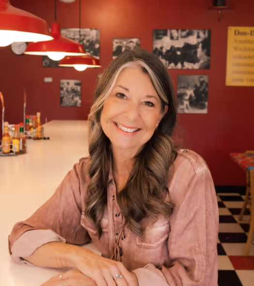 Smiling middle-aged woman with long wavy hair sitting at a diner counter with red walls and vintage photos behind her.