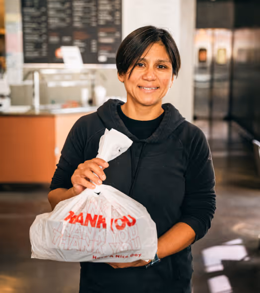 Smiling person holding a white plastic takeout bag with red 'Thank You' text inside a restaurant.
