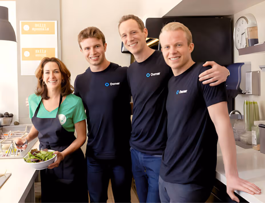 Four smiling employees in a kitchen, one woman holding a bowl of salad and three men wearing matching black shirts with 'Owner' logos.