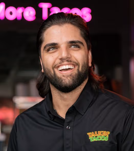 Smiling man with a beard wearing a black shirt with a colorful Talkin' Tacos logo.