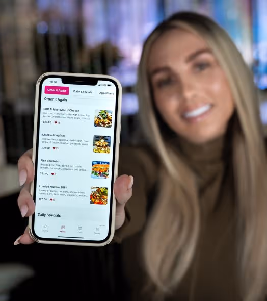 Woman showing a smartphone screen with a food delivery app menu displaying items like BBQ Brisket Mac N Cheese, Chicken & Waffle, Fish Sandwich, and Loaded Nachos.