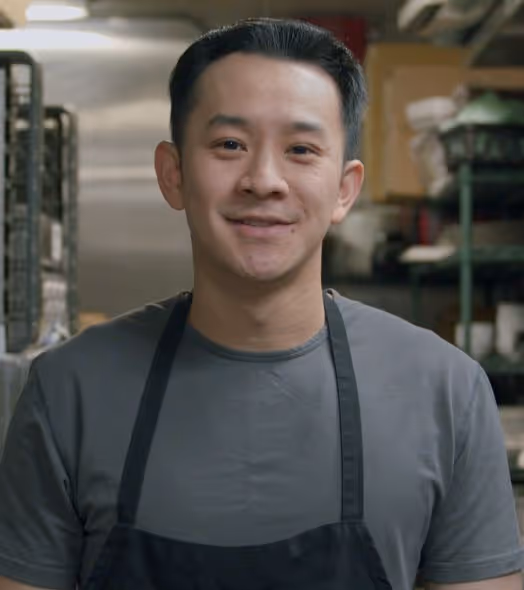 Smiling man wearing a black apron and gray shirt standing in a kitchen with shelves and racks in the background.