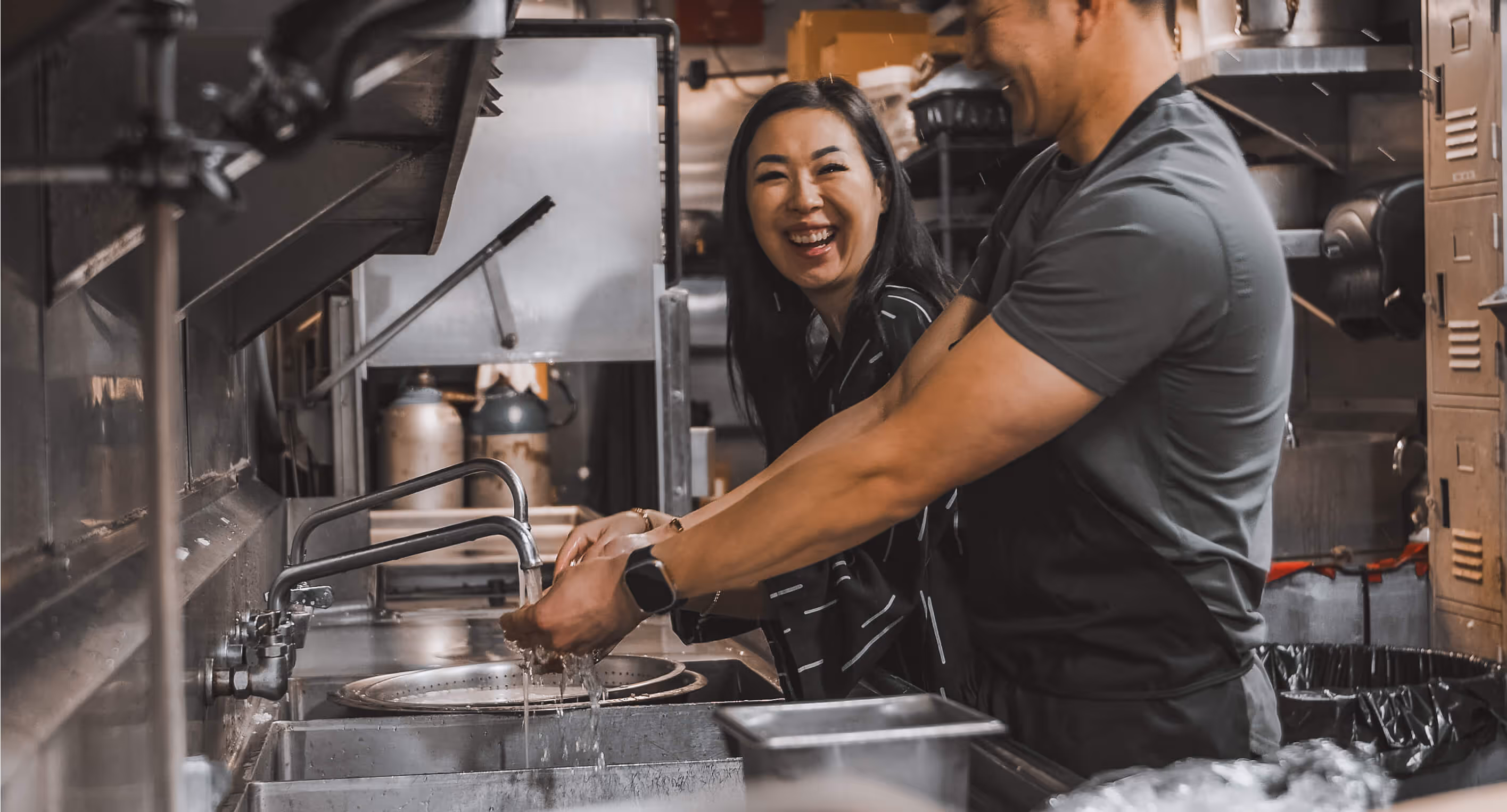 Man and woman laughing together while washing hands in a commercial kitchen sink.