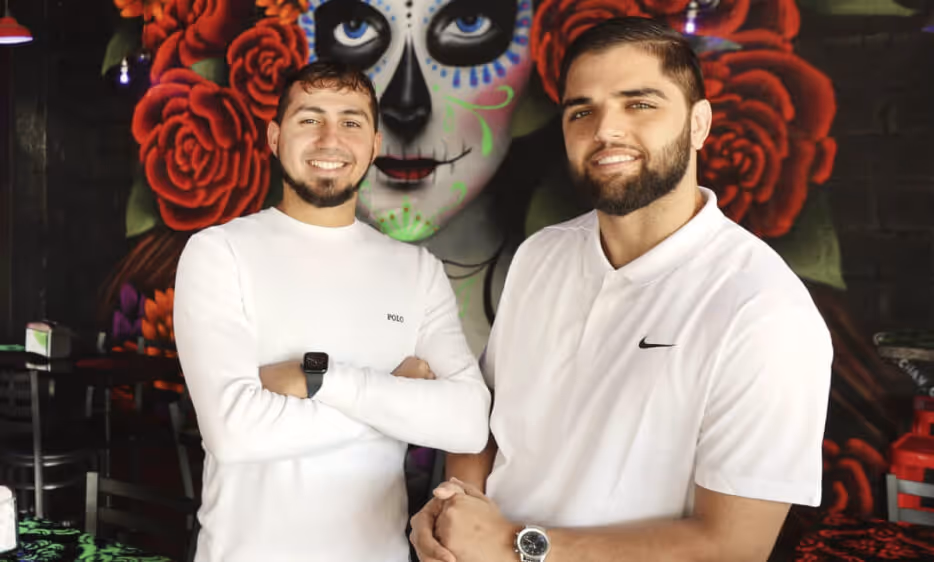 Two men smiling, wearing white shirts, standing in front of a mural of a face painted like a sugar skull with red roses.