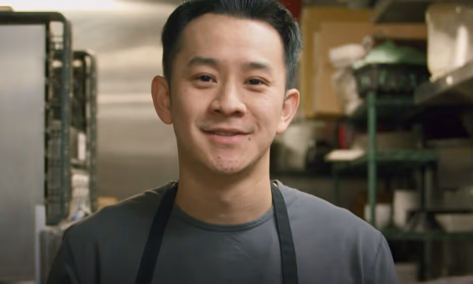 Young man wearing a gray shirt and black apron smiling in a kitchen setting.
