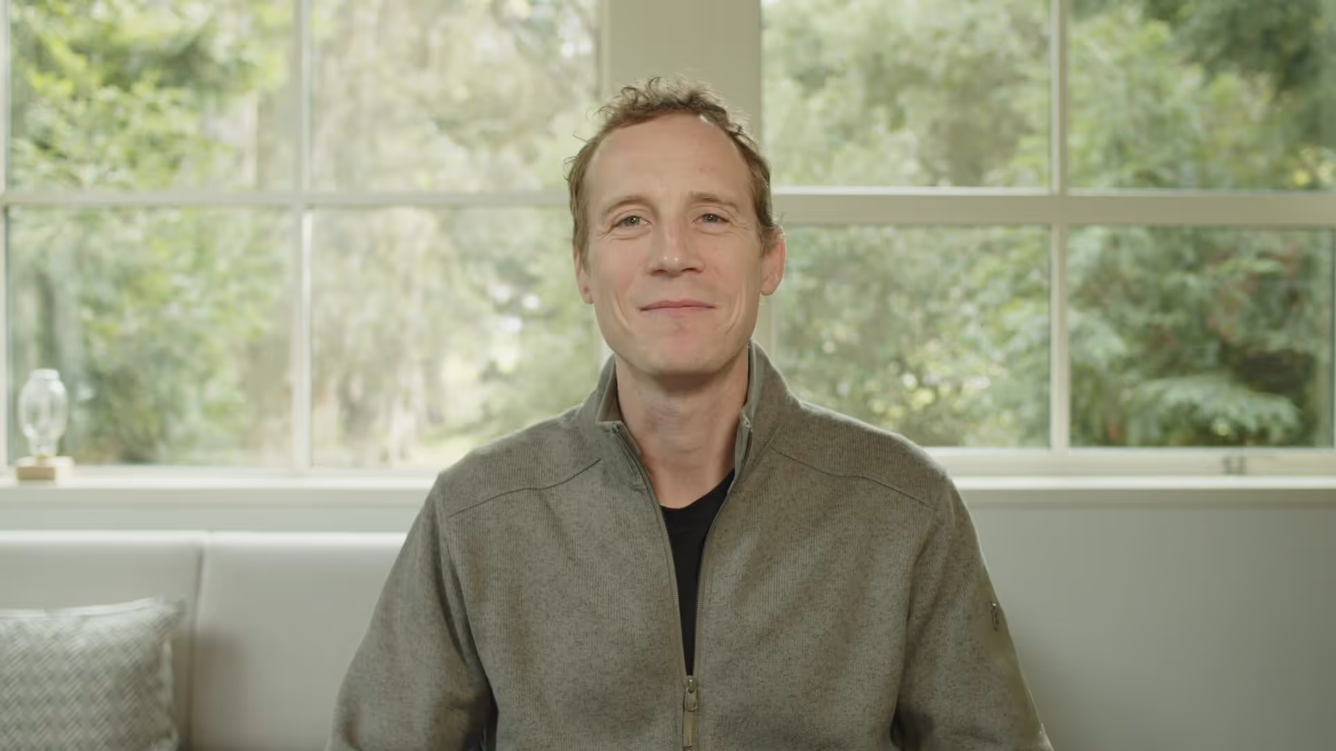 Smiling man wearing a gray zip-up jacket sitting on a white couch in front of a large window with green trees outside.