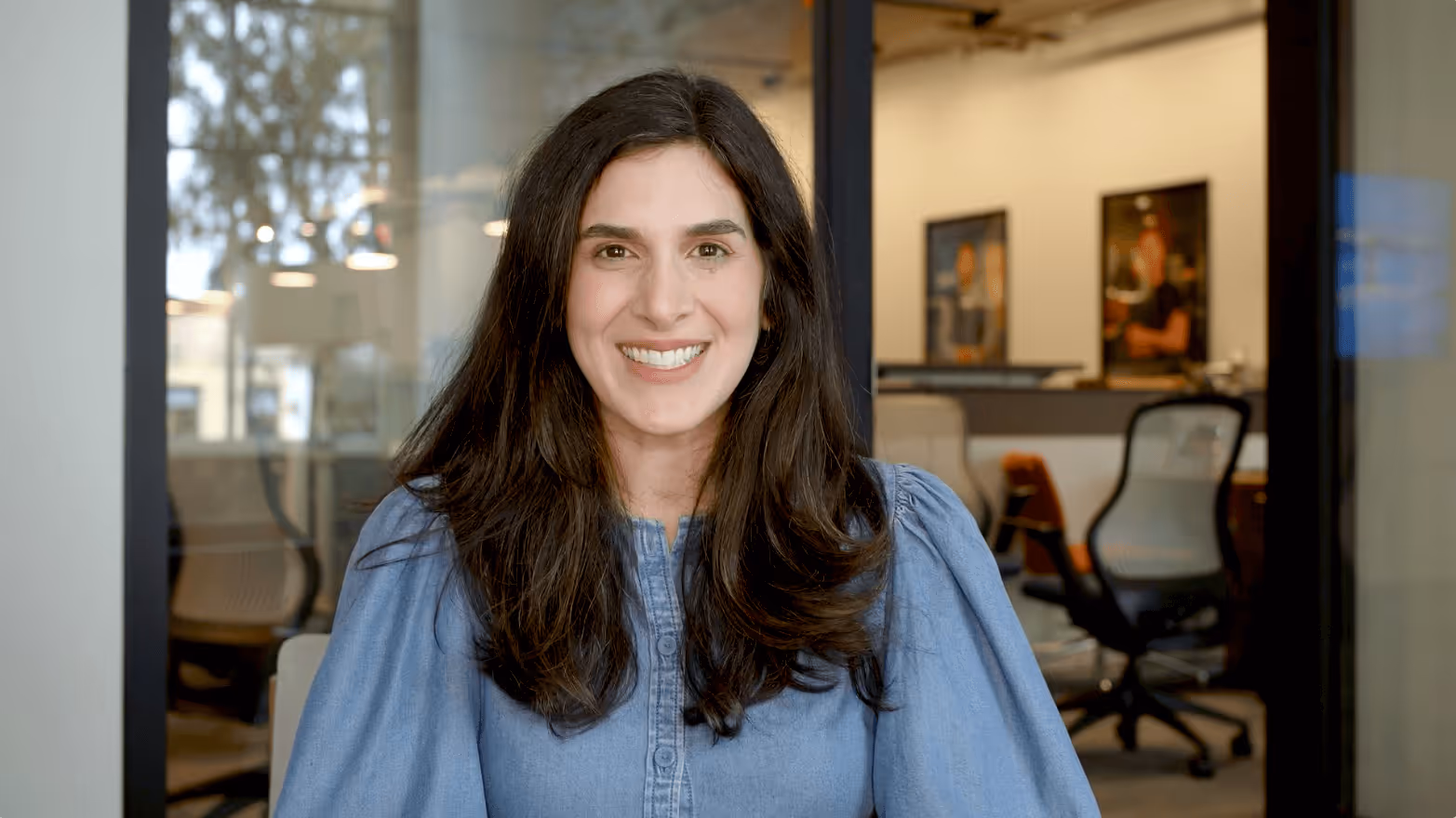 Smiling woman with long dark hair wearing a blue blouse sitting in a modern office.
