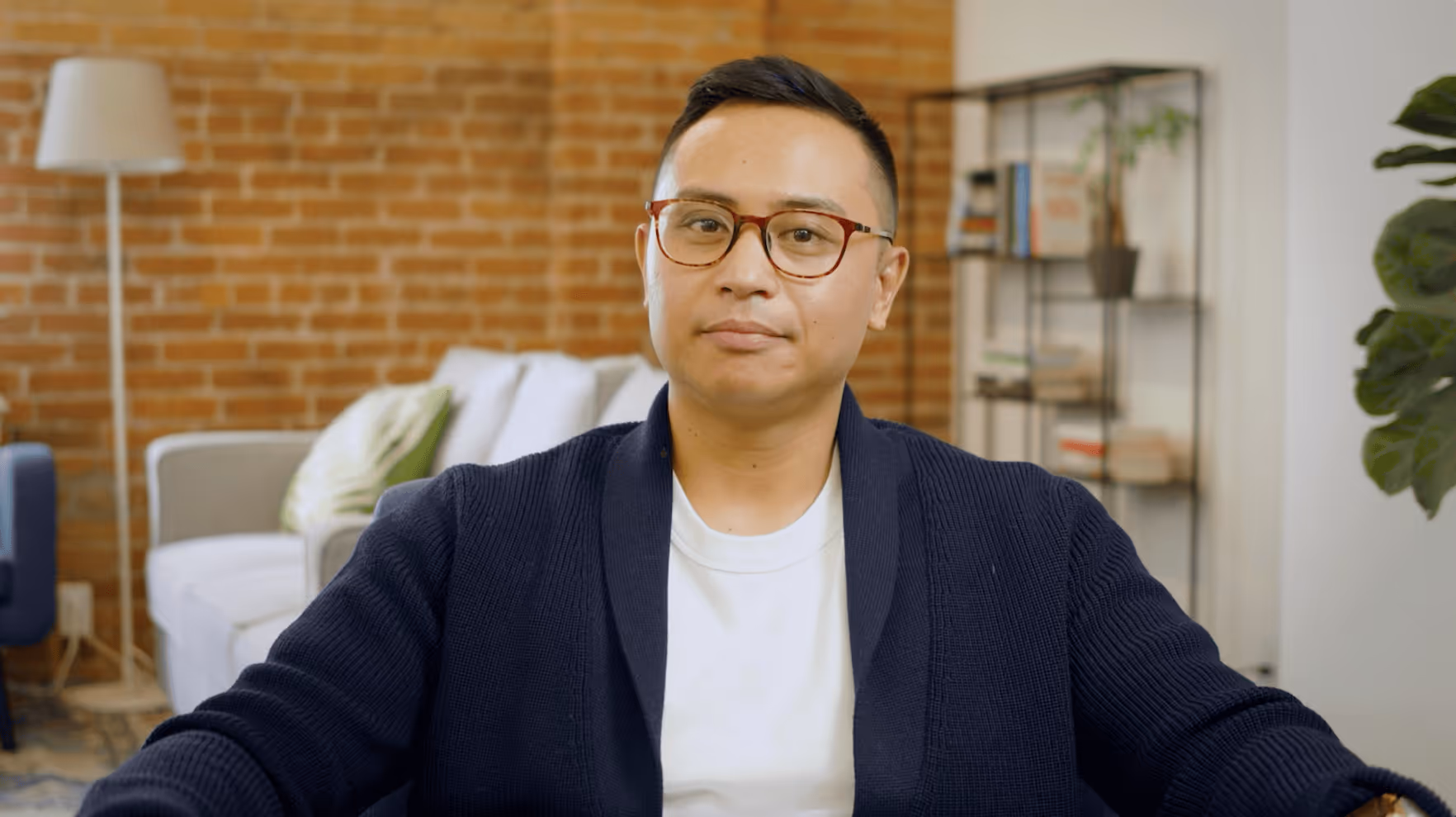Man wearing glasses, a white shirt, and a navy cardigan sitting in a cozy room with exposed brick walls and bookshelves.