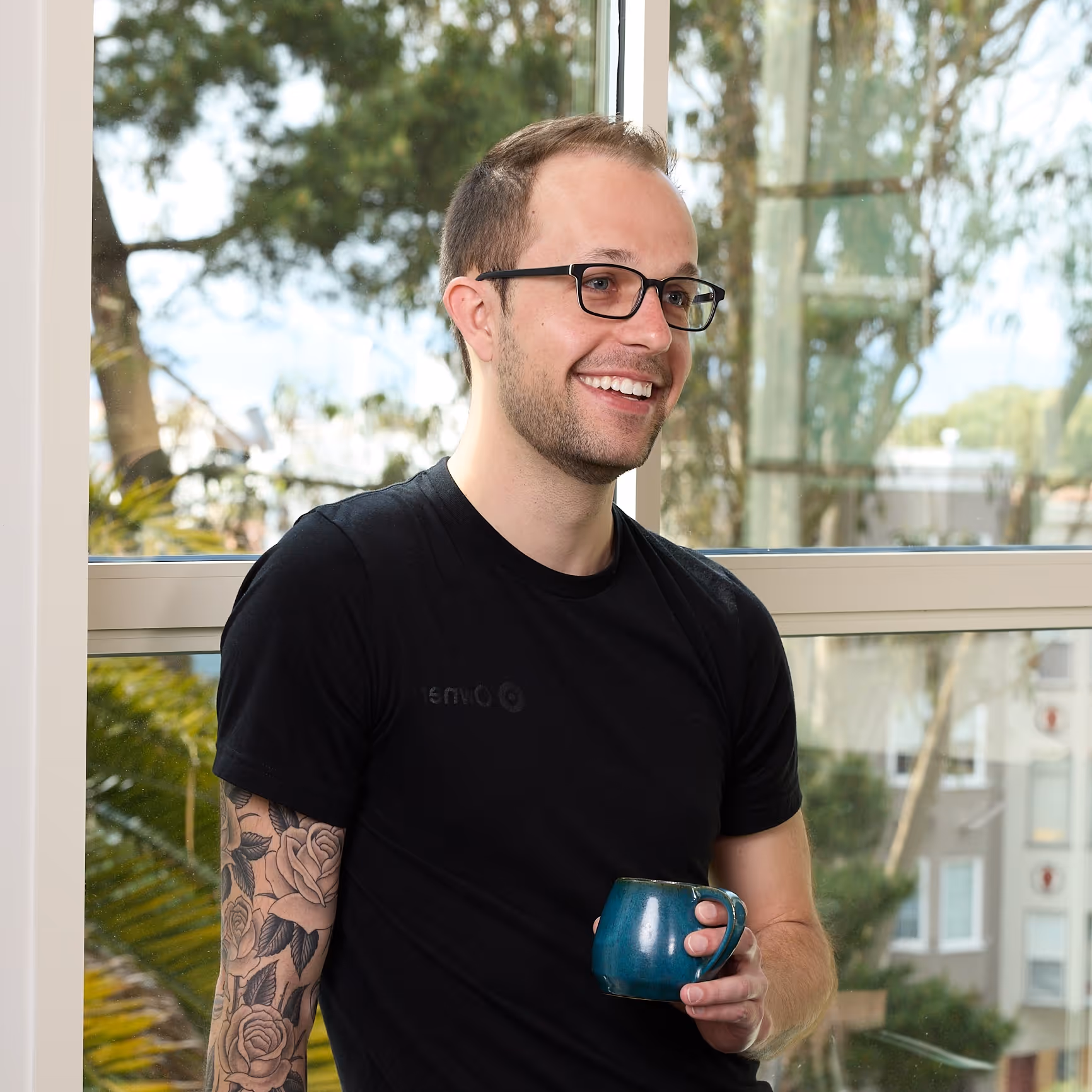 Smiling man with glasses and a floral tattoo on his arm, holding a blue ceramic mug while standing in front of a large window.