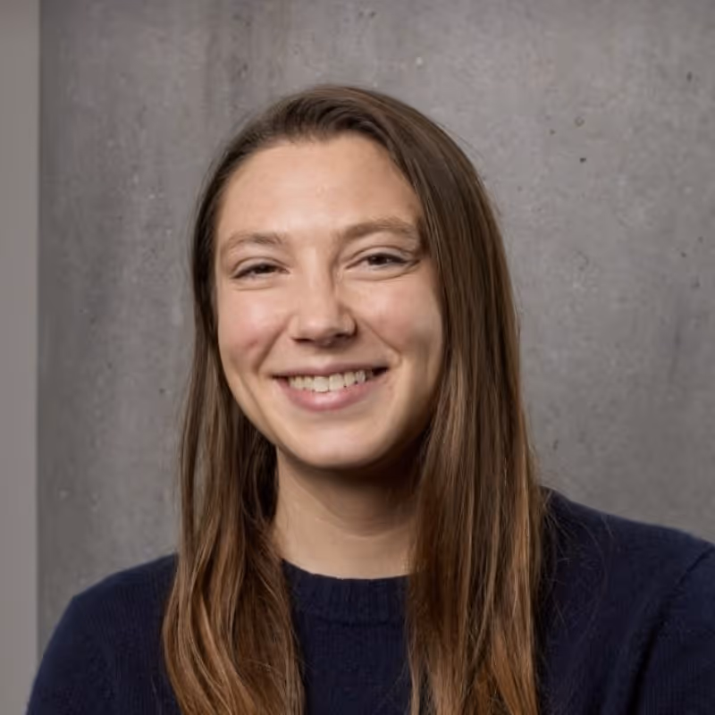 Smiling young woman with long brown hair wearing a dark sweater against a gray wall background.