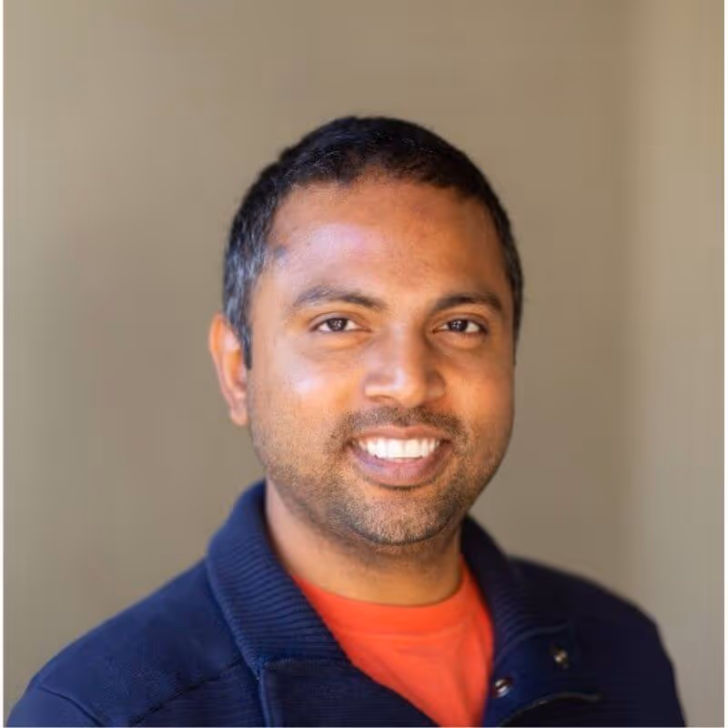 Smiling man with short black hair wearing a navy jacket and red shirt against a beige background.