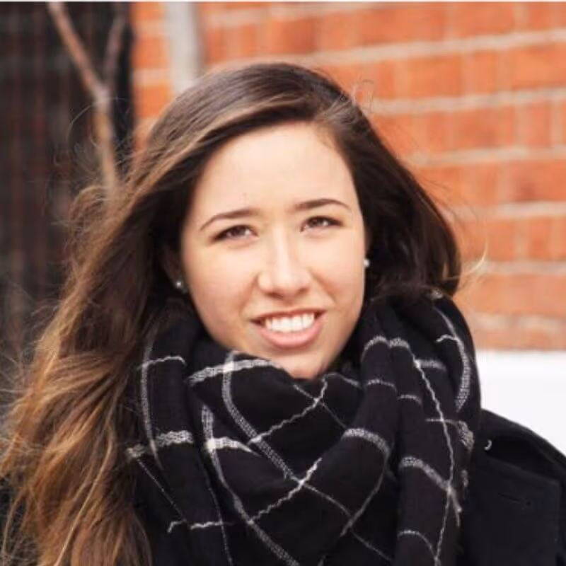 Young woman with long brown hair smiling, wearing a black and white checkered scarf and black coat, standing in front of a brick wall.