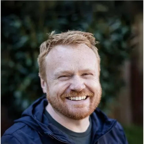 Smiling red-haired man with beard wearing a dark jacket, outdoors with blurred greenery background.