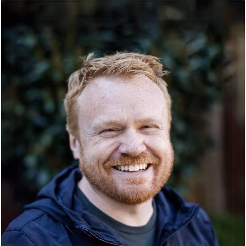 Smiling red-haired man with beard wearing a dark jacket, outdoors with blurred greenery background.