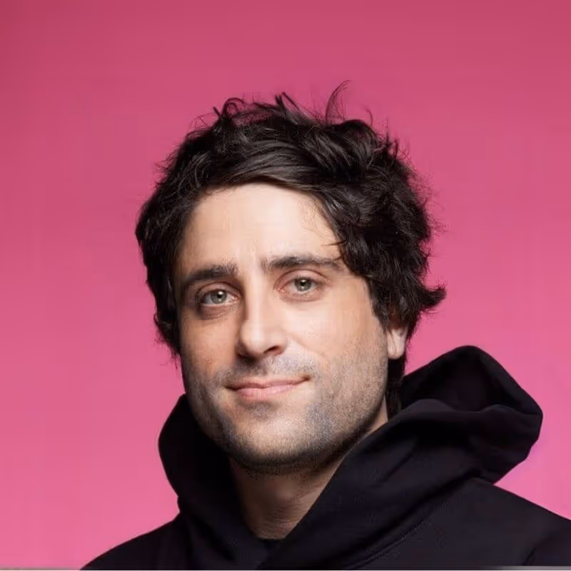 Portrait of a man with dark curly hair wearing a black hoodie against a pink background.