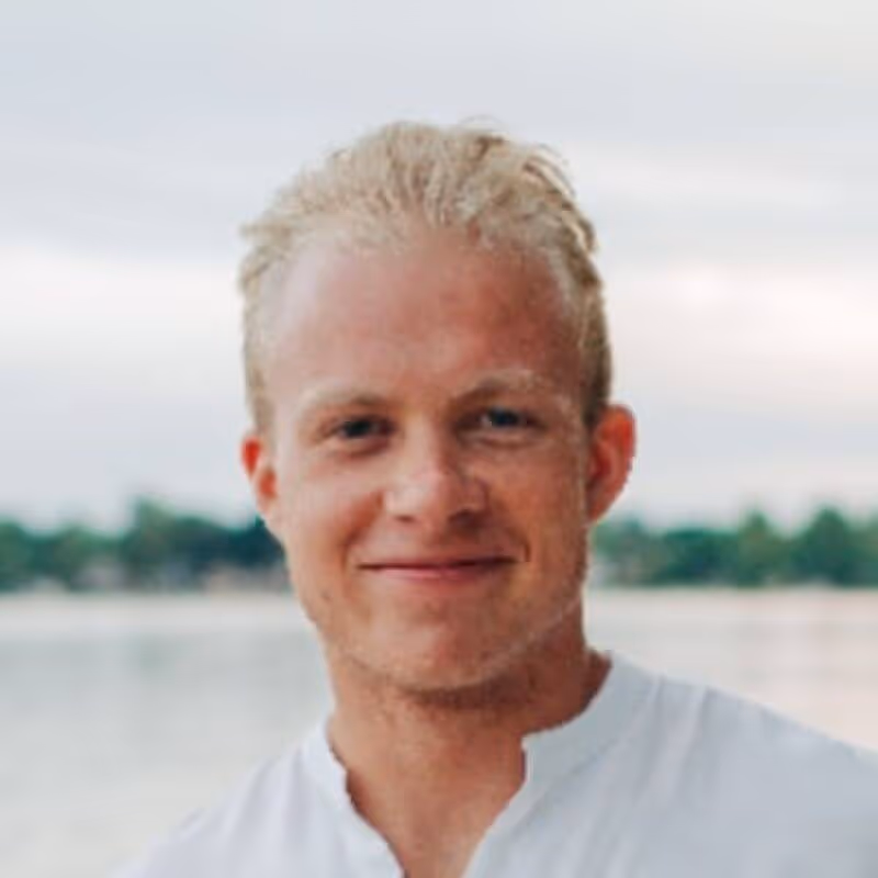 Smiling young man with short blond hair wearing a white shirt, standing outdoors near a body of water.