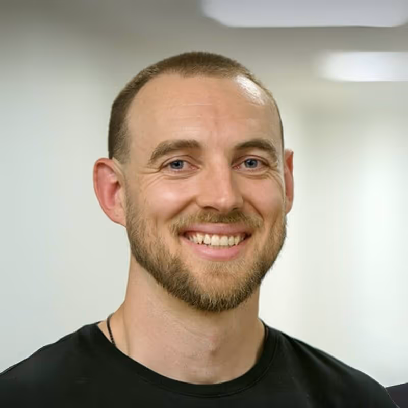 Smiling man with short hair and beard wearing a black shirt against a light background.