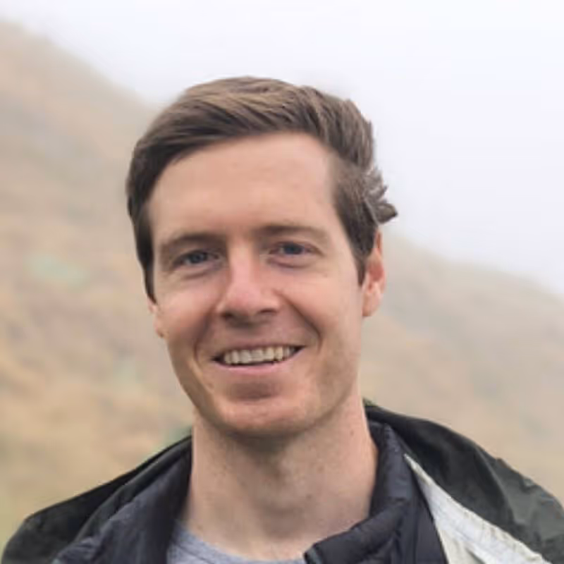 Portrait of a smiling young man with light brown hair wearing a jacket outdoors with a blurred natural background.