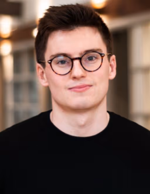 Young man with short dark hair and round glasses wearing a black shirt in an indoor setting.