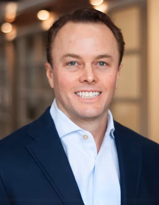 Smiling man with short brown hair wearing a white dress shirt and dark blazer in a softly lit indoor setting.