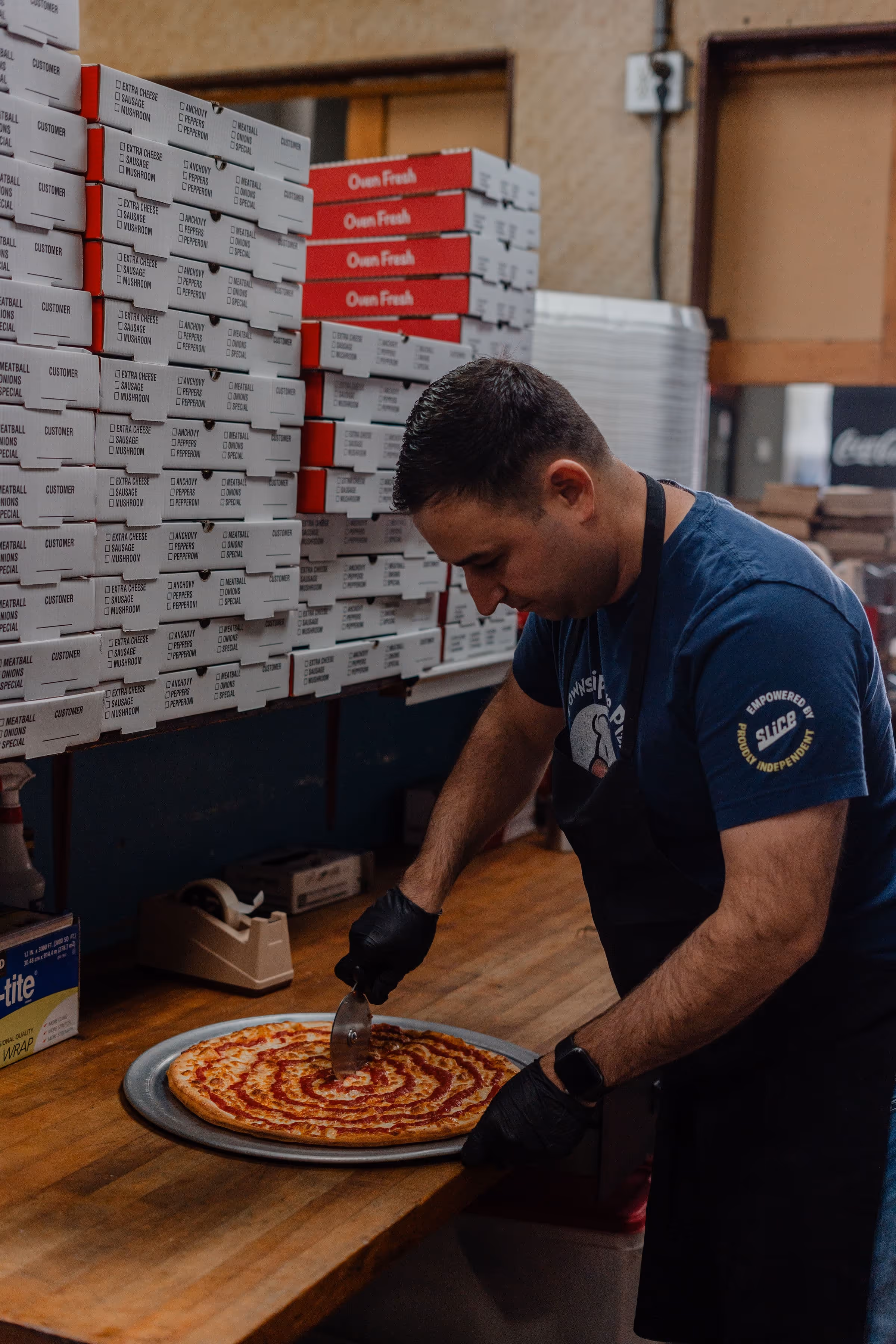 Man wearing black gloves and apron using a pizza cutter to slice a large pizza with spiral sauce on a wooden counter next to stacked pizza boxes.