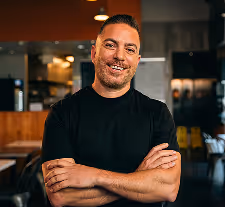 Smiling man with short hair and beard wearing a black t-shirt, standing with arms crossed in a warmly lit indoor setting.