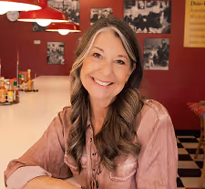 Smiling middle-aged woman with long brown hair sitting at a restaurant counter with red walls and black-and-white photos in the background.