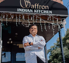 Man standing confidently with arms crossed in front of Saffron Indian Kitchen restaurant entrance.