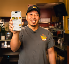 Smiling man in a baseball cap and t-shirt holding up a smartphone displaying an app inside a restaurant.