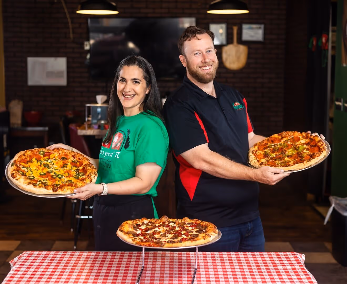 Smiling man and woman holding pizzas in a restaurant with a third pizza on a stand on a checkered tablecloth in front of them.