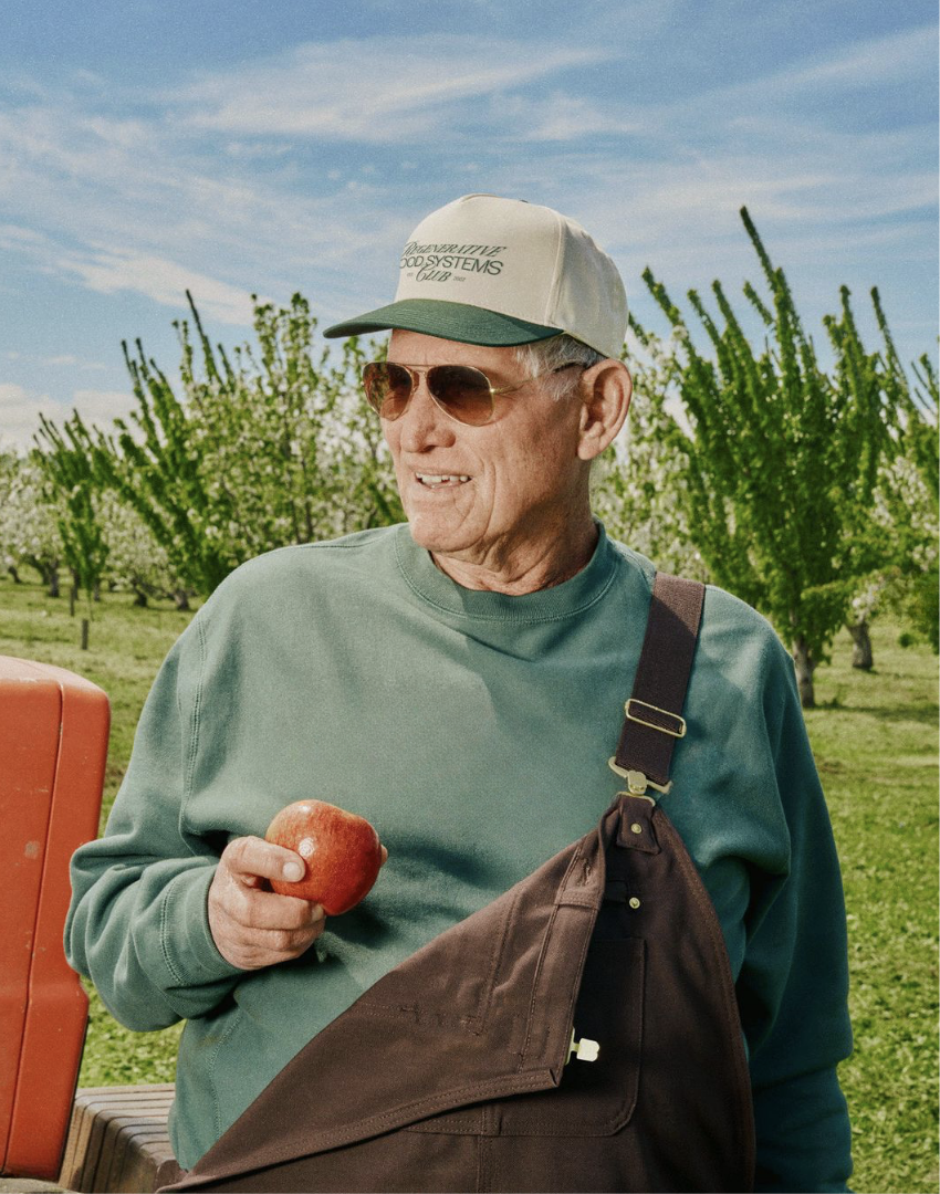 An older man wearing a cap and sunglasses stands in an orchard holding an apple and smiling.