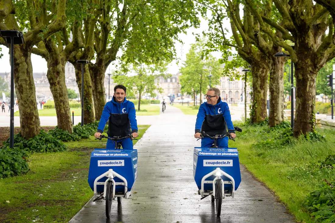 Deux plombiers "Coup de clé" en uniforme bleu sur des vélos cargo dans un parc urbain à Bordeaux