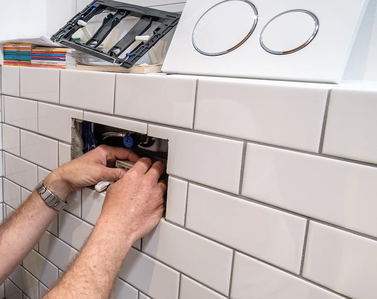 Plombier réparant un WC encastré, panneau de chasse retiré et outils visibles, mur carrelé dans salle de bain