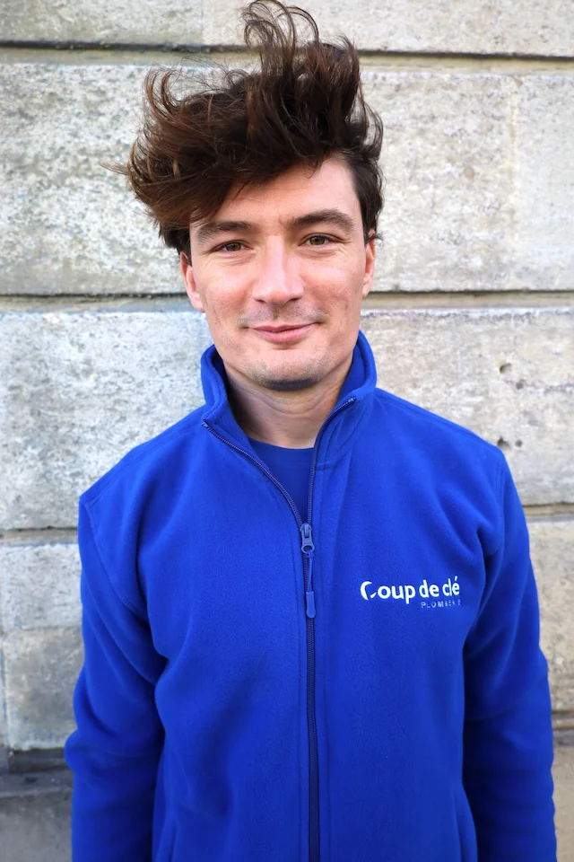 Romain, membre de l’équipe Coup de clé, en veste polaire bleue avec le logo de l’entreprise, devant un mur en pierre