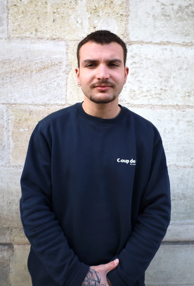 Tristan, membre de l’équipe Coup de clé, en sweat-shirt bleu marine avec le logo de l’entreprise, bras croisés devant un mur en pierre
