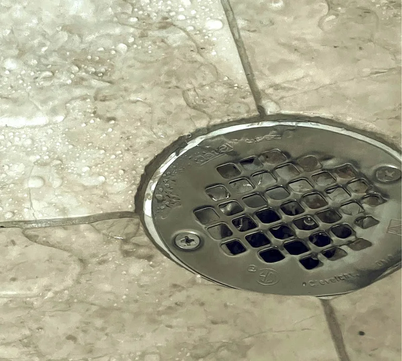 Jet d'eau clair tombant dans un drain de sol en métal sur un carrelage beige mouillé.