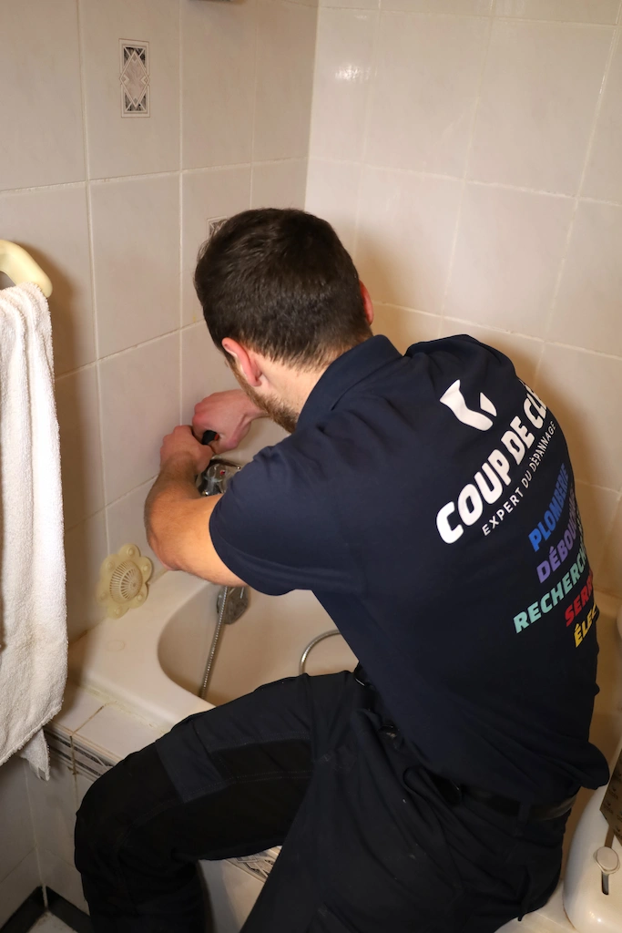 Un plombier en uniforme bleu foncé effectue des réparations dans une baignoire carrelée.