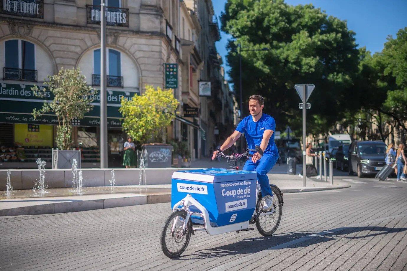 Homme en tenue bleue conduisant un vélo de livraison avec une grande caisse bleue portant le logo Coup de clé sur une rue ensoleillée.