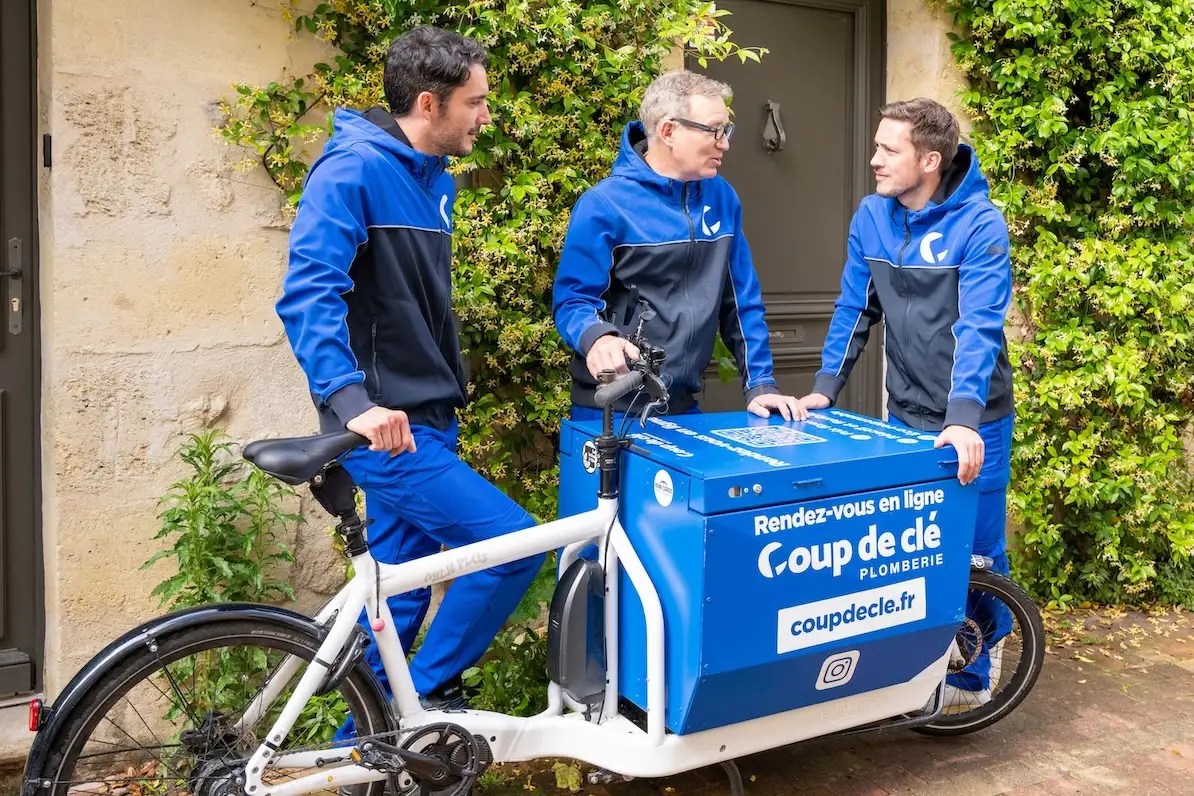 Trois plombiers en vêtements de travail bleus discutent devant un vélo-cargo blanc avec une boîte bleue marquée « Coup de clé » devant un immeuble.